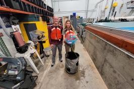 两名UNH学生研究人员将他们赢得的海洋可再生能源设备放在波浪箱前. 他们穿着红色的救生衣.